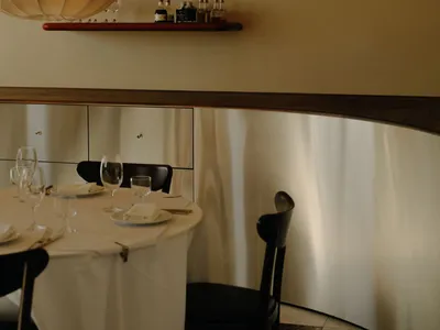 Elegant dining area featuring a curved walnut wall, built-in floating shelves, black dining chairs, and a round table set with white linens under a large pendant lamp.