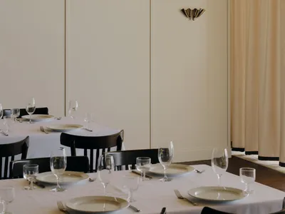 Elegant restaurant dining room featuring dark walnut chairs, white tablecloths, and tall cream curtains with a decorative brass wall sconce.
