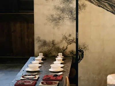 長型深色石桌上陳列白色瓷茶杯與紅色托盤上的彩色串珠珠寶，背景為大型中國水墨屏風，繪有蒼勁老樹，呈現極簡侘寂風室內空間。