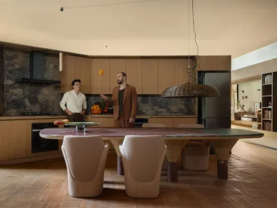 Modern open-plan kitchen and dining area with a large curved travertine dining table, sculptural wooden legs, beige dining chairs, light wood cabinetry, and a dramatic stone feature wall.
