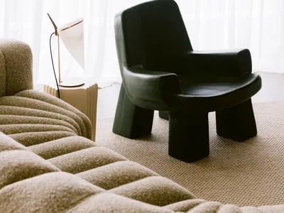 Modern living room corner featuring a black sculptural armchair, a beige boucle sofa, a white table lamp on a fluted side table, and floor-to-ceiling sheer white curtains.