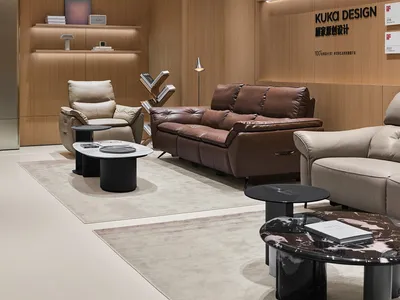 Modern luxury living room display with brown leather sofa, cream leather armchairs, black marble coffee tables, and warm wood panel walls with integrated lighting.