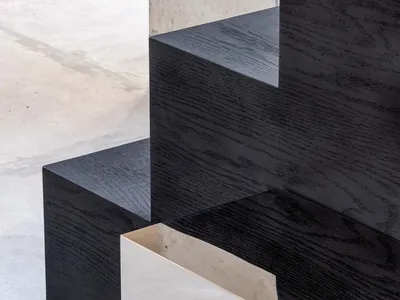 Close-up view of a modern staircase featuring thick black oak wooden treads with visible grain, cantilevered over a light gray polished concrete floor, with a textured concrete wall in the background.