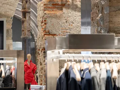 Interior view of a fashion boutique featuring rough exposed brick walls, stone arches, modern stainless steel clothing racks, and a mannequin wearing a bright red dress.