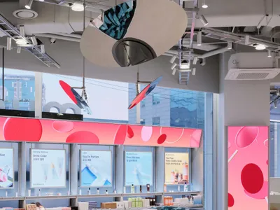 Modern beauty store interior with a large curved grey counter, colorful product displays, digital pink LED wall graphics, and hanging sculptural light fixtures.