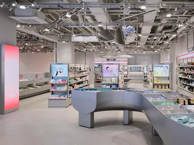Interior view of a modern cosmetics and skincare store featuring a large curved grey display island, product shelving, digital advertisements, and exposed ceiling infrastructure.