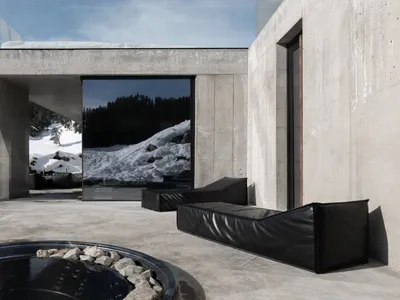 Modern concrete villa exterior with black leather daybeds, circular hot tub, and large glass windows overlooking snowy mountain landscape.