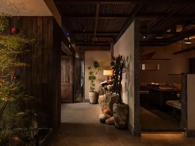 日式餐廳昏暗的走廊，深色木牆、質感石柱、盆栽植物，以及可看到溫暖用餐區與長椅座位的視野。