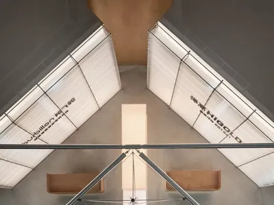 Top-down architectural view of a modern concrete atrium with two suspended brown leather benches, steel cable system, and large angled skylights with white blinds.