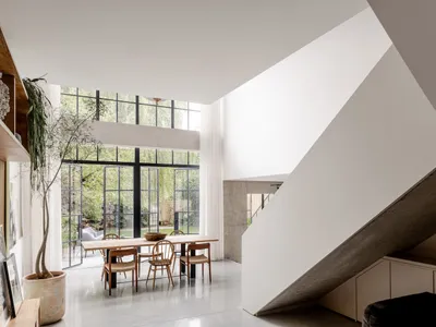 Modern double-height dining room featuring a large wooden table, woven dining chairs, concrete staircase, and expansive black-framed windows overlooking a garden.