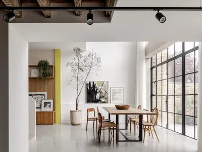 現代工業風格 loft 餐廳，長型橡木餐桌搭配多款木製餐椅、大型盆栽樹木、白磚牆，以及引入充足自然光的大型黑色格窗。
