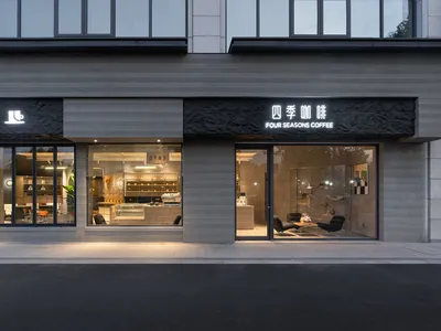 四季咖啡店街景，灰色紋理混凝土現代立面、大片玻璃窗展示內部，以及黃昏時分亮起的黑色石材招牌。