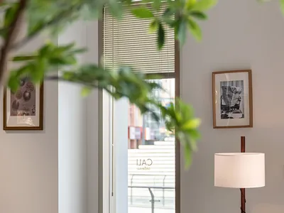 Modern cafe interior featuring round wooden tables, walnut bentwood chairs, a tall floor lamp, framed art, and a large window with blinds letting in natural light, partially framed by foreground plant leaves.