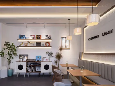 Modern cafe interior with wooden dining tables, grey upholstered banquette seating, white brick wall with backlit signage, floating wooden shelves displaying vinyl records, and warm pendant lighting.