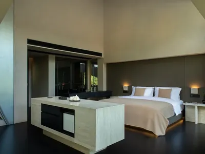Modern minimalist bedroom featuring a large travertine stone island, low platform bed with beige bedding, dark polished floor, and warm wall sconces under high angled ceilings.