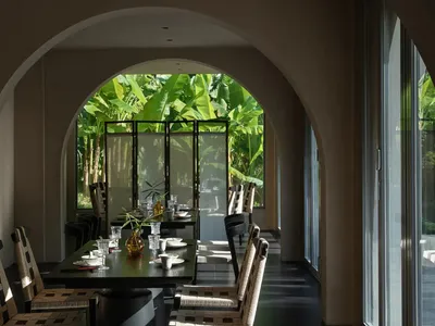 Modern dining room featuring a large arched doorway that frames a tropical garden with banana trees, a dark dining table set with white ceramics, and chairs with black and cream woven rattan backs.