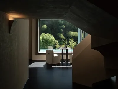 Modern minimalist living space featuring raw concrete architecture, a white armchair, round side table, and large window revealing a dense forest view.