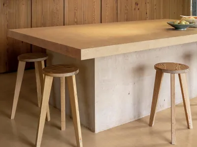 Modern minimalist kitchen island made of concrete and wood, paired with two large woven rattan pendant lights and three wooden stools against a vertical wood-paneled wall.