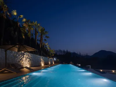 A luxurious infinity pool glowing with underwater lights at night, bordered by a textured natural stone wall, palm trees, and lounge chairs overlooking dark mountains.