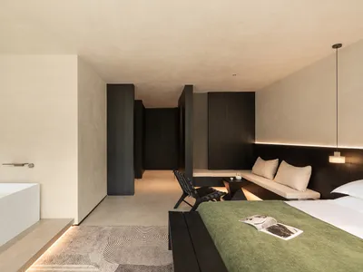Modern minimalist bedroom featuring a freestanding white bathtub, olive green bedspread, dark wood accents, and micro-cement walls with integrated lighting.