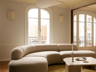 A spacious Parisian living room featuring a large curved off-white boucle sectional sofa on a round chartreuse rug, with three sculptural white pendant lights hanging from a ceiling with ornate plasterwork, arched French windows, and herringbone wood flooring.