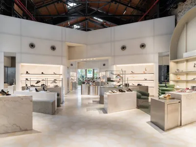 Modern minimalist shoe store interior with white marble display islands, integrated LED lighting, green velvet seating, and exposed industrial ceiling with skylights.