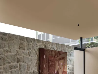 穿著咖啡色西裝的男子坐在酒紅絲絨與格紋混搭的模組沙發上，前方是大面積天然石牆與大型畫作，地板鋪設綠色地毯並有室內植物。