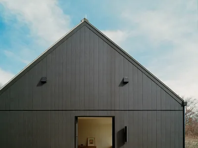 Modern black barn house with large gable roof, open sliding door revealing a bedroom interior, wooden deck with railing, and a single red Adirondack chair under a blue sky.