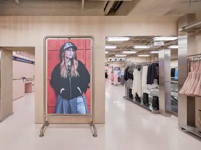 Modern fashion boutique interior featuring light wood paneling, a large digital portrait on a metal stand, stainless steel accents, and clothing racks in the background.