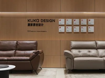 Modern showroom interior with a brown leather sofa on the left and a beige leather sofa on the right, set against a backdrop of light wood paneling with KUKA DESIGN branding and multiple iF and Red Dot design awards displayed.