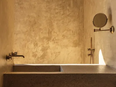 Modern minimalist bathroom featuring a concrete shower-bath, textured plaster walls, and warm brass fixtures with a round magnifying mirror.