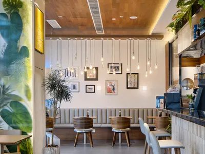Modern cafe interior with large tropical leaf mural on the left, multiple wooden pendant lights hanging from a wooden ceiling, upholstered banquette seating, and round wooden tables with white chairs.