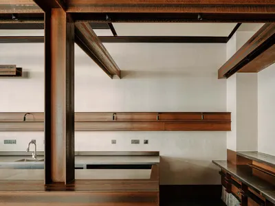 Modern kitchen interior featuring a raised platform made of weathered corten steel, massive exposed dark steel beams, minimalist white walls, and a stone base.