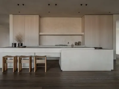 現代極簡風廚房，大型混凝土中島結合水槽與爐具，三張曲線木製吧台椅，淺木色系統櫃與深色木地板。