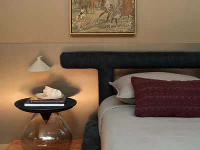 Modern bedroom corner with dark boucle upholstered headboard, terracotta knit throw, black side table with glass base, and framed antique tapestry with jewel embellishments on a neutral wall.