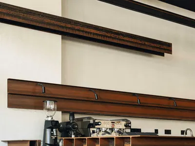 Modern industrial coffee station featuring a counter constructed from stacked weathered corten steel I-beams, with professional espresso machines and coffee grinders against a white wall with dark steel beams overhead.