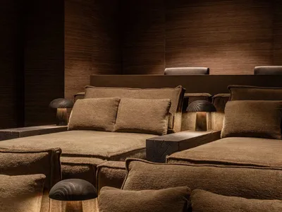 Low-profile modular sofa upholstered in rich brown boucle fabric, arranged with multiple textured cushions, set against dark wood panel walls and illuminated by three black mushroom table lamps.