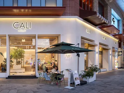 Modern white cafe exterior at night with green outdoor seating, large glass windows, and warm interior lighting showing patrons inside.
