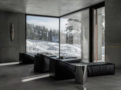 Modern minimalist living room with raw concrete walls, large floor-to-ceiling windows overlooking a snowy mountain forest, black leather modular sofas, and brushed metal side tables.