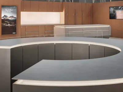 Modern luxury boutique interior featuring a large curved grey reception island in the foreground, warm oak cabinetry and backlit display wall in the background, with large windows covered by horizontal blinds.