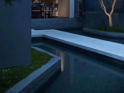 Night view of a modern concrete house courtyard with a linear water feature, moss garden, small tree, and warmly lit indoor dining area visible through large openings.