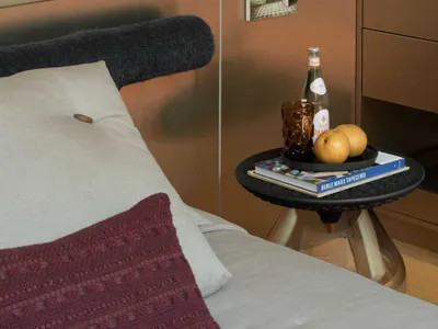 Modern bedroom corner featuring a bed with white and burgundy pillows, a black textured side table with brass base, a minimalist white floor lamp, and warm metallic wall panels.