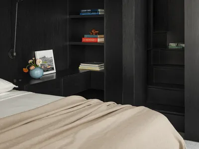 Modern dark wood loft bedroom featuring a raised sleeping platform, built-in bookshelves, a wall-mounted reading lamp, and neutral bedding with soft beige tones.