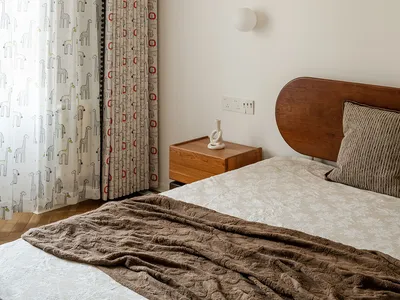 Child's bedroom corner showing a wooden bed with textured pillow, brown throw blanket, oak nightstand with white sculpture, and playful giraffe and geometric patterned curtains allowing natural light.