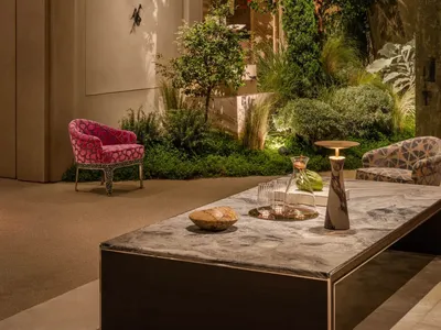 Modern lobby lounge featuring a large indoor garden with lush trees and plants, a marble coffee table, a vibrant pink patterned armchair, and a geometric patterned sofa under warm ambient lighting.