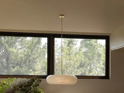 Modern dining room featuring a round travertine table, four dark wood woven-seat chairs, a large alabaster pendant lamp, and a large window overlooking green trees.
