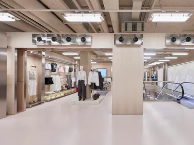 Wide view of a modern minimalist clothing boutique interior with concrete pillars, light birch wood partitions, white floors, and exposed ceiling infrastructure.