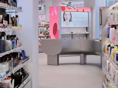 Modern cosmetics store interior with curved grey central island, pink digital display walls, and neatly organized product shelves under bright track lighting.