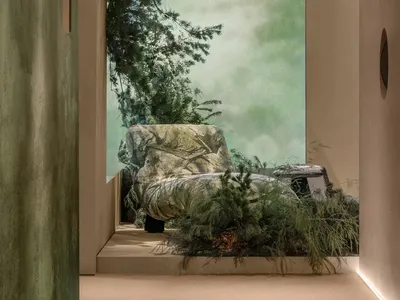 Modern biophilic interior featuring a stone-like armchair surrounded by lush evergreen plants, with soft natural light filtering through a large opening and curved plaster architecture.