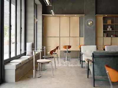 Modern cafe interior featuring concrete walls, light wood cabinetry, built-in window seating, round glass tables, and a mix of upholstered and molded wood chairs in green, orange, and neutral tones.